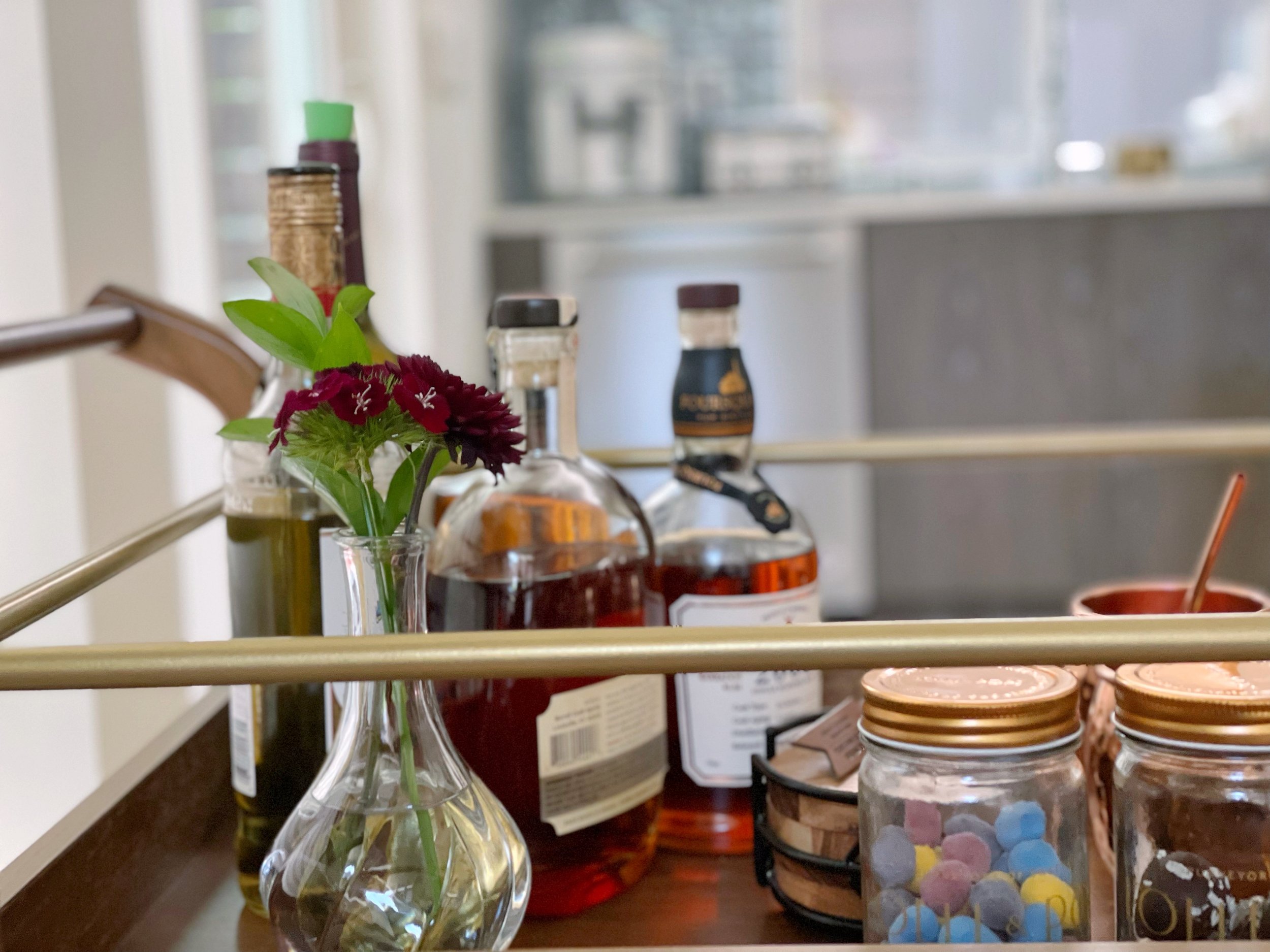 A bar cart with liquor bottles, a small vase of flowers, and jars with colored candies.