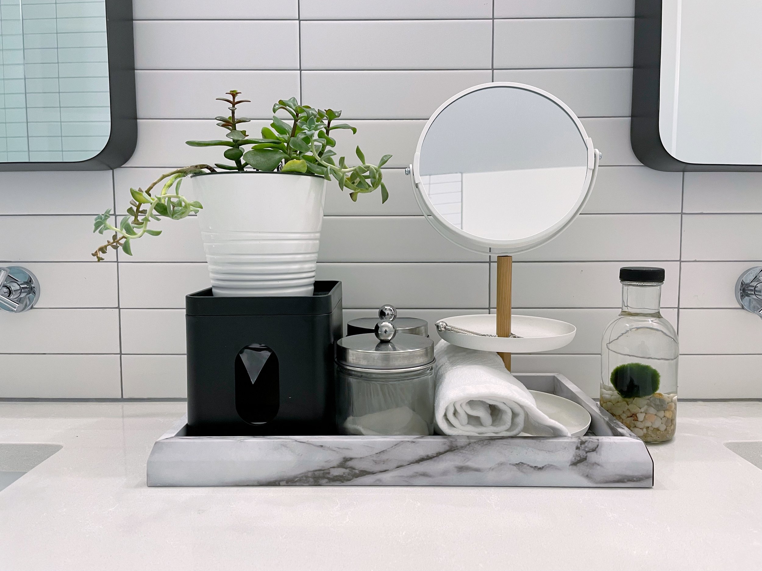 Bathroom countertop with a potted plant, mirror, rolled towel, jar, and bottle with green marimo moss ball on a marble tray.