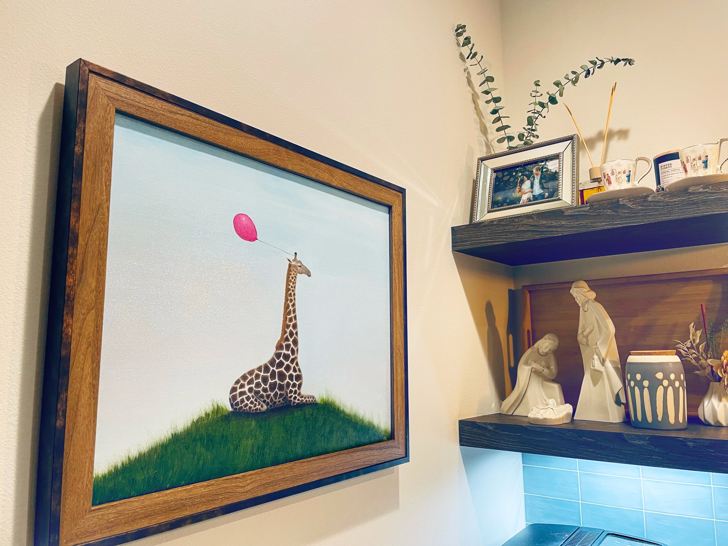 Framed painting of a giraffe with a red balloon, next to shelves with a nativity scene and decorative items.