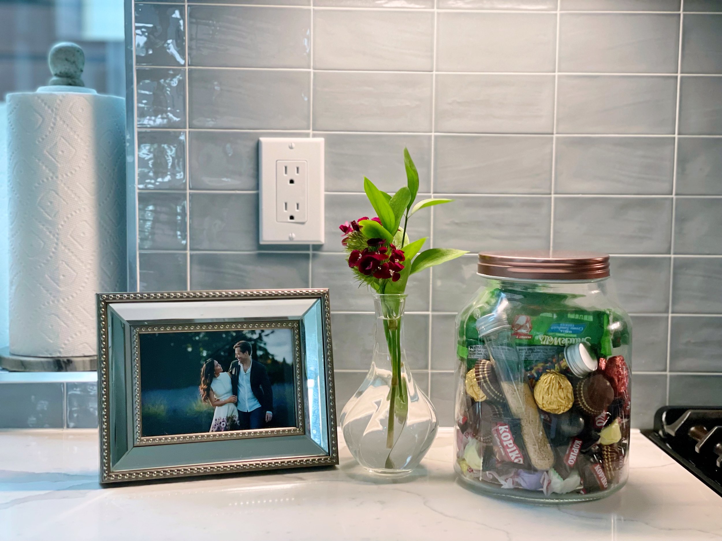 Kitchen countertop with a gray tile backsplash, a framed photo, a vase with red flowers, and a jar of candy.