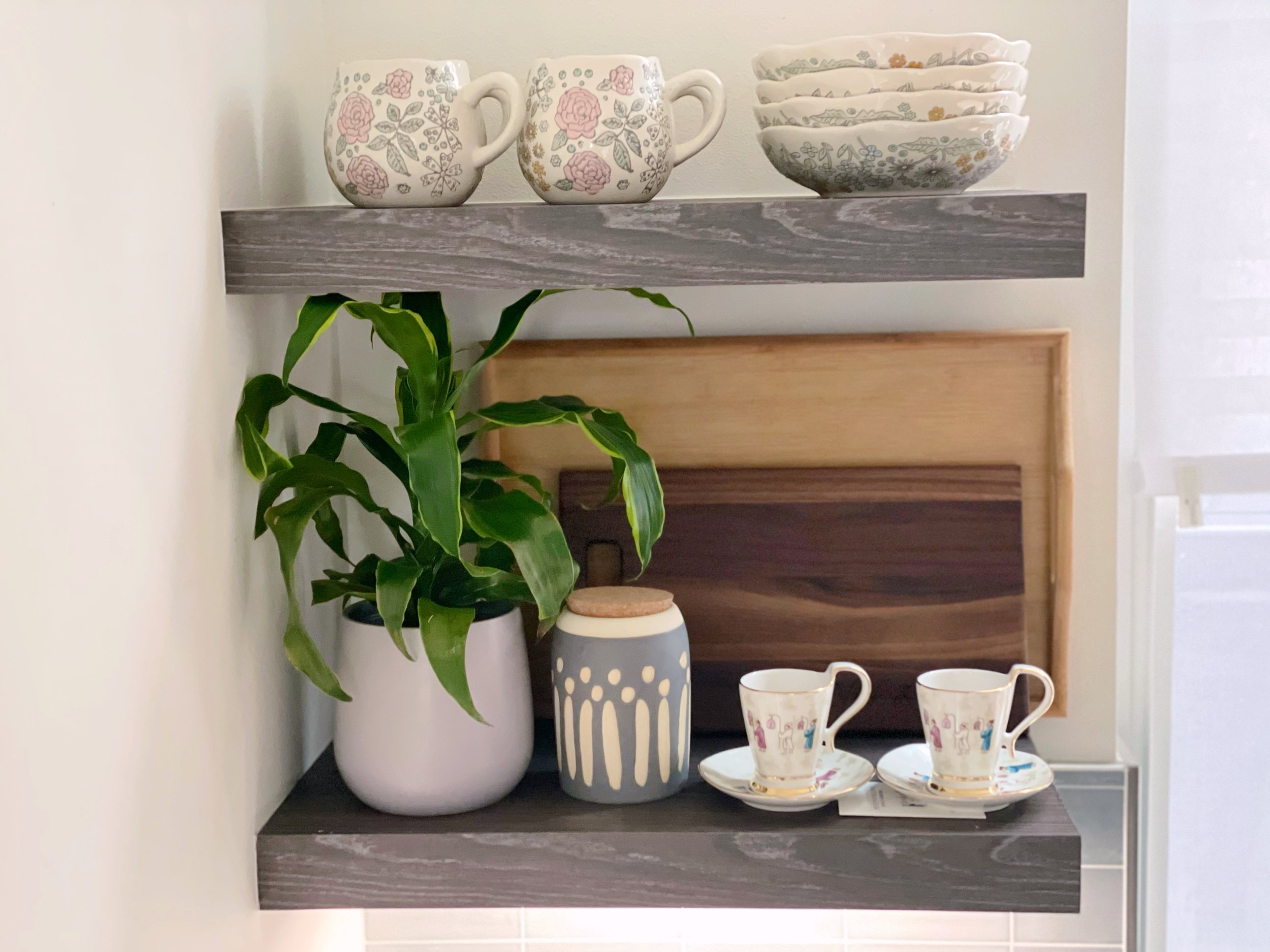 Kitchen shelf with mugs, bowls, a potted plant, wooden cutting boards, and teacups with saucers.