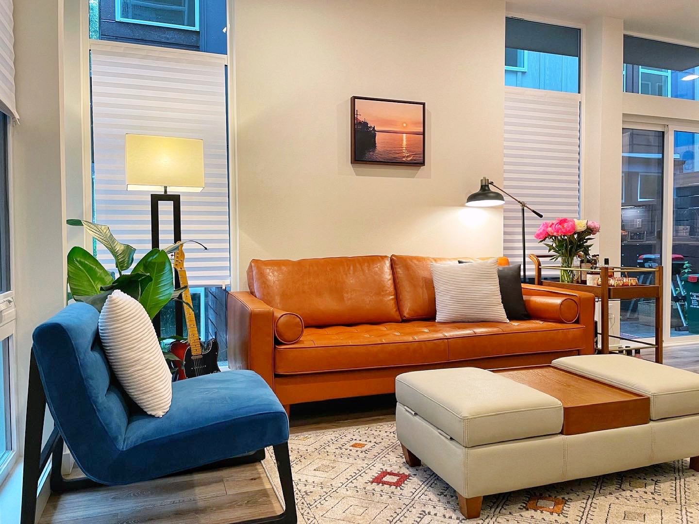 Modern living room with a tan leather sofa, blue accent chair, large potted plant, floor lamp, and a guitar.