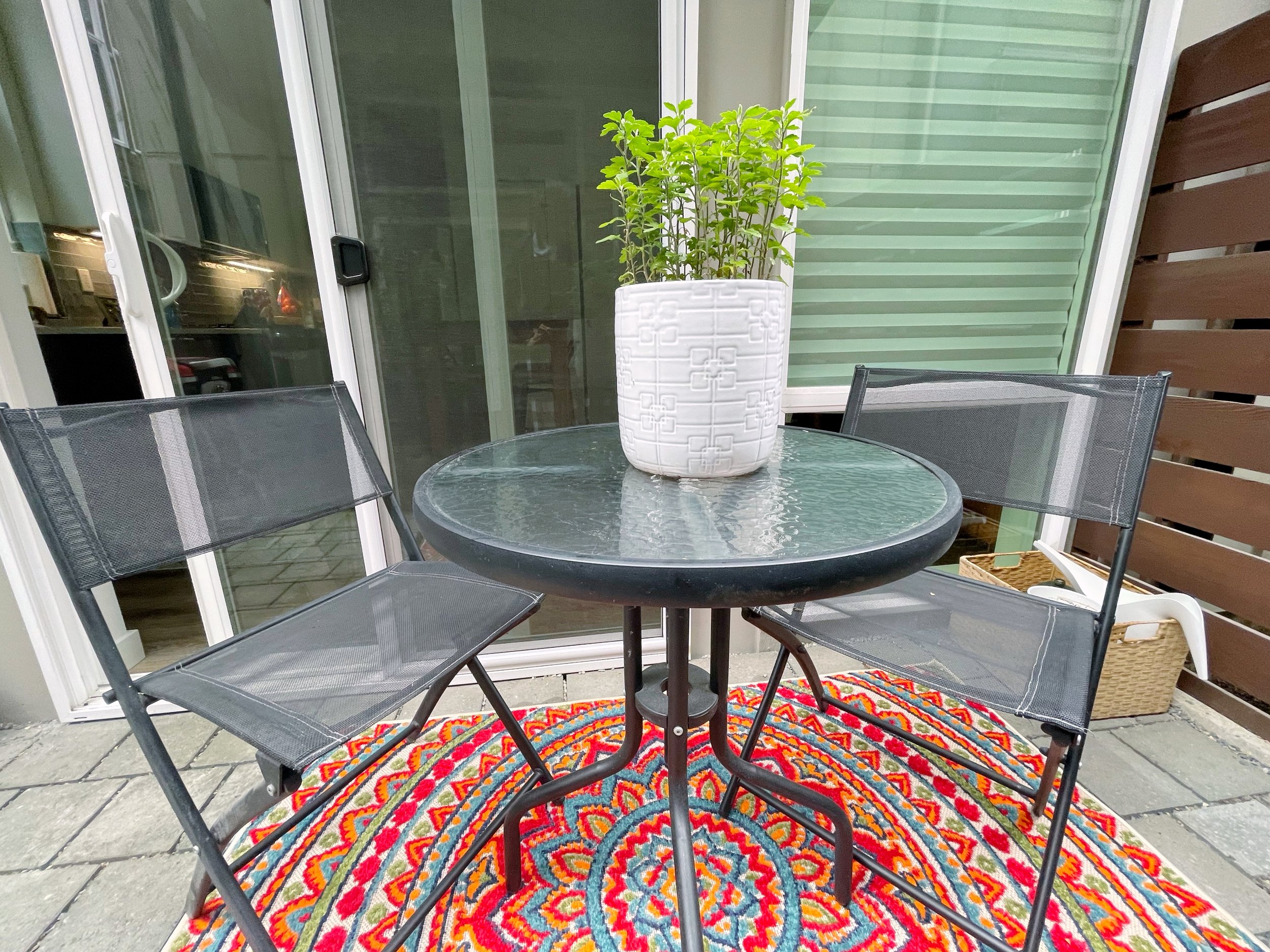 Patio set with a glass table and two mesh chairs on a colorful round rug, in front of sliding glass doors.