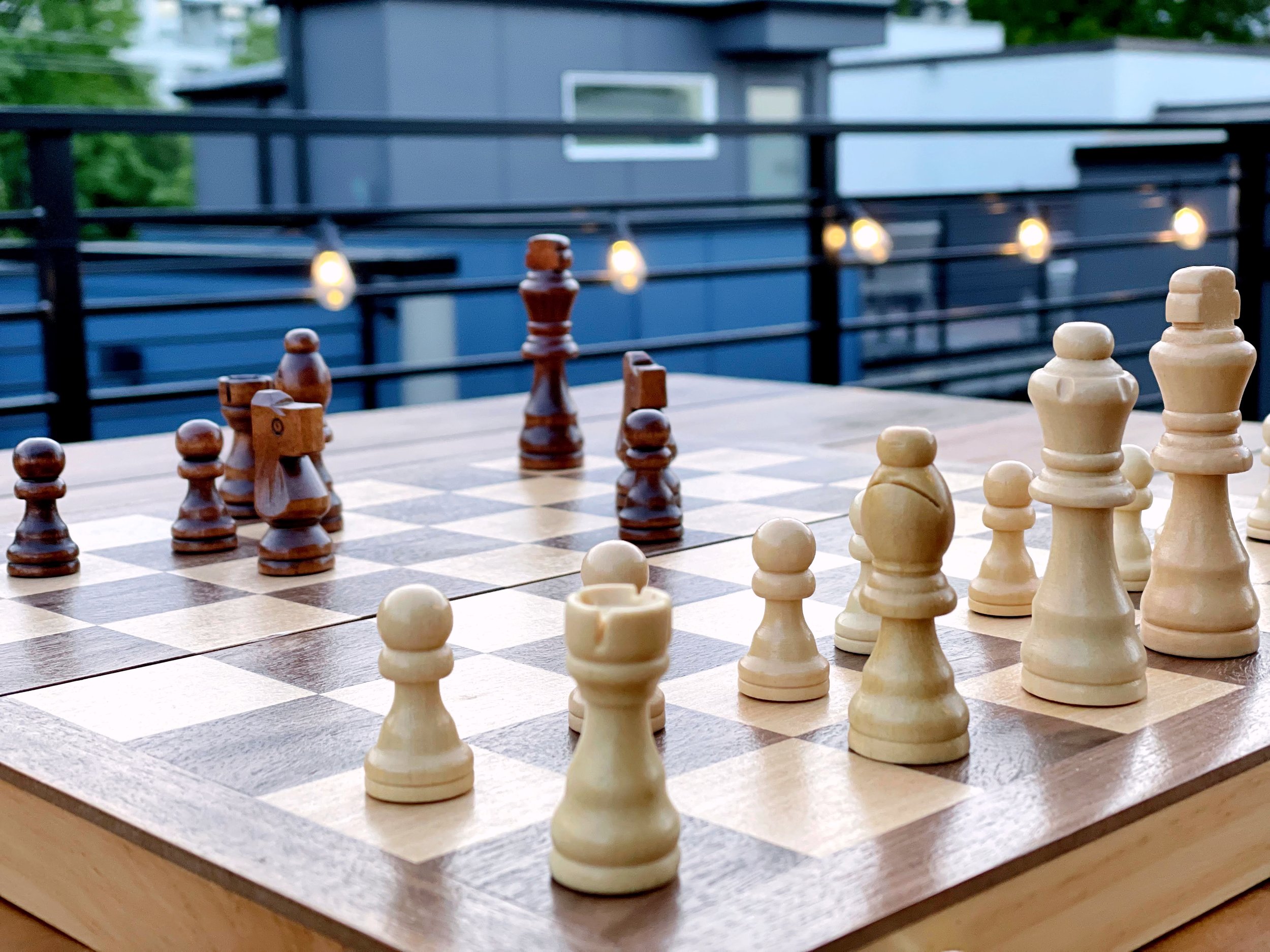 Outdoor chess game set up on a wooden board with black and white pieces, light bulbs and building in the background.