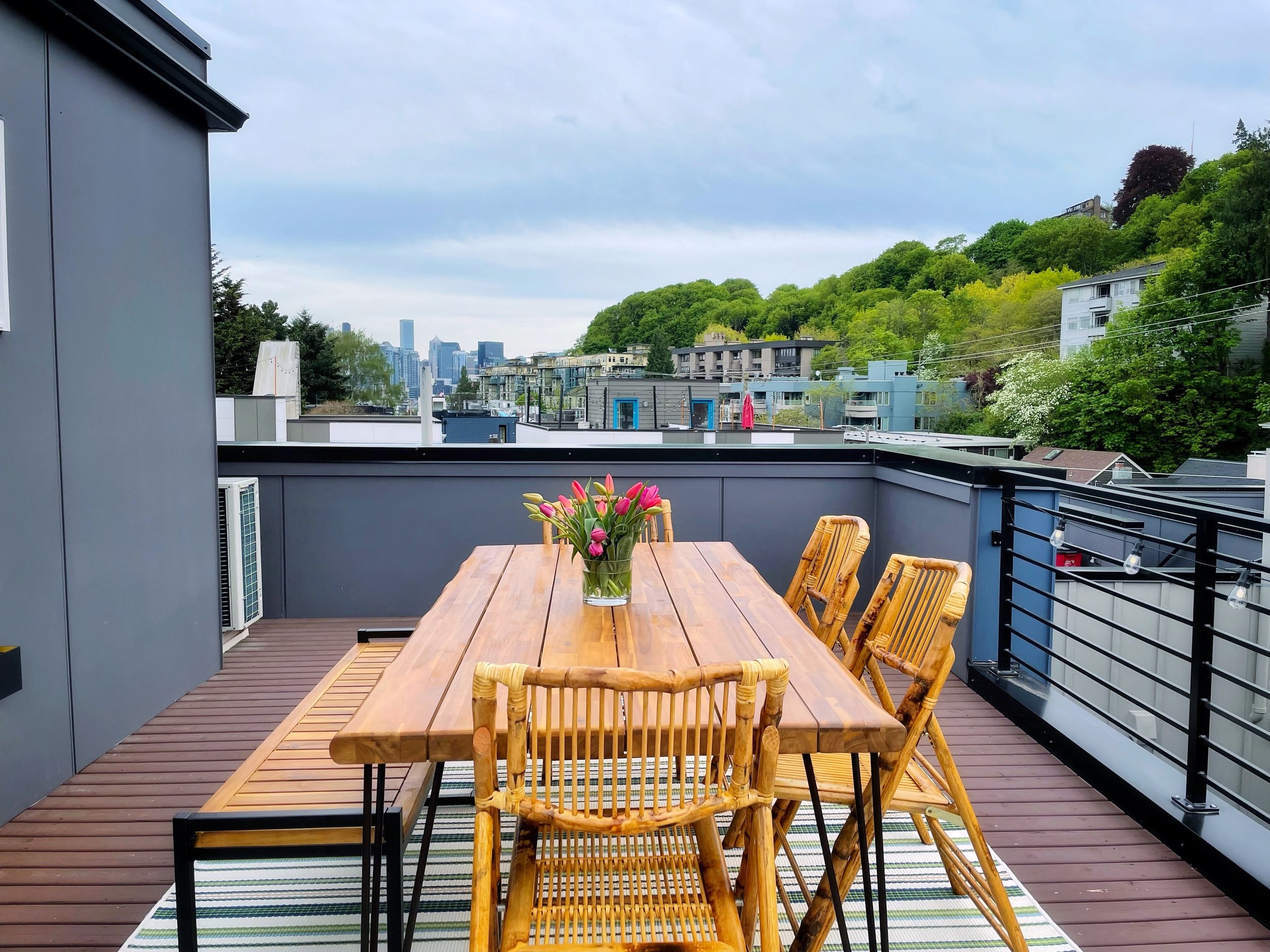Rooftop patio with a wooden dining table and chairs, vase with flowers, overlooking a city skyline.
