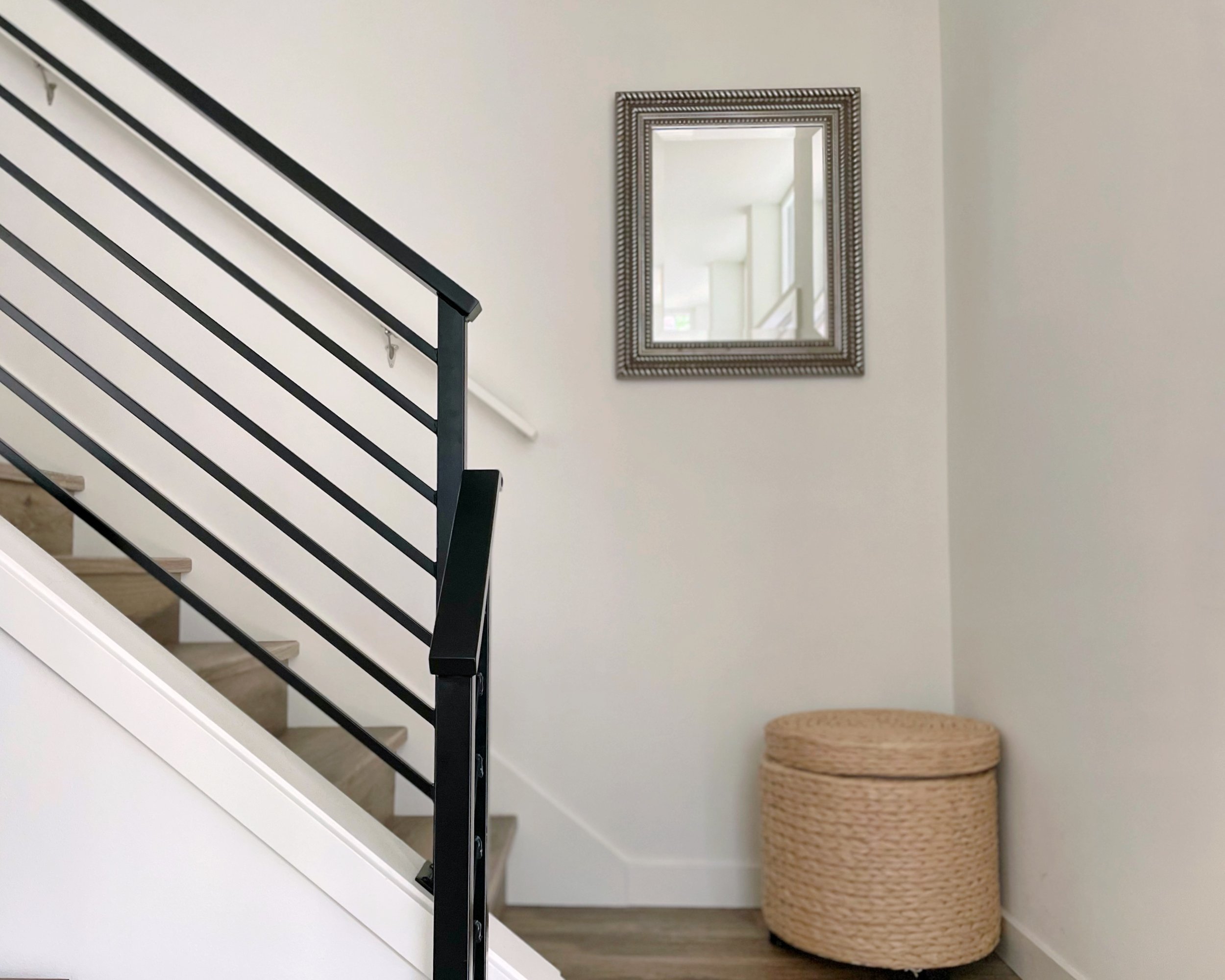 Modern staircase with metal railing, a woven ottoman, and a decorative wall mirror.